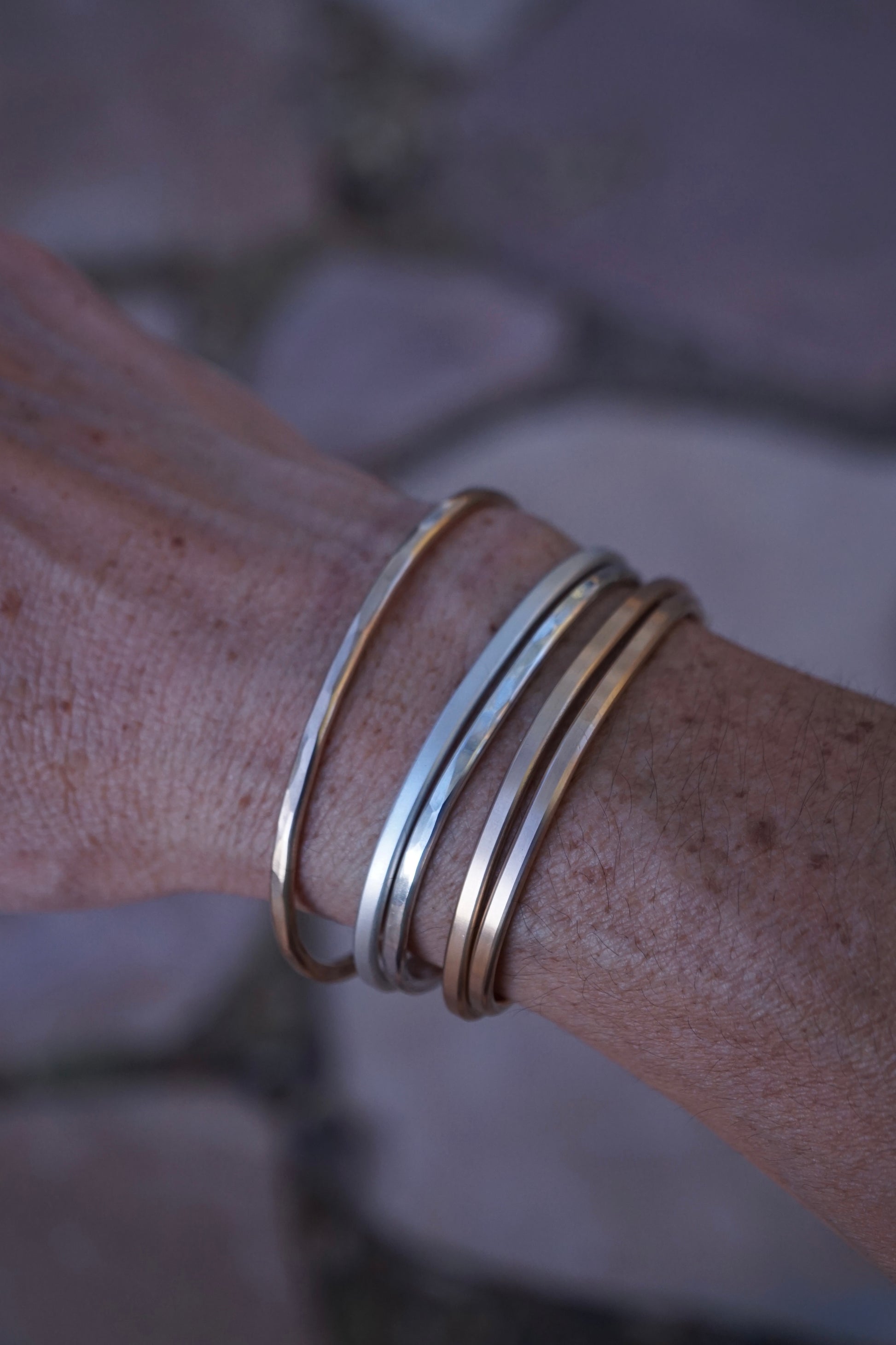 Close-up of a wrist wearing multiple gold bracelets against a blurred natural background